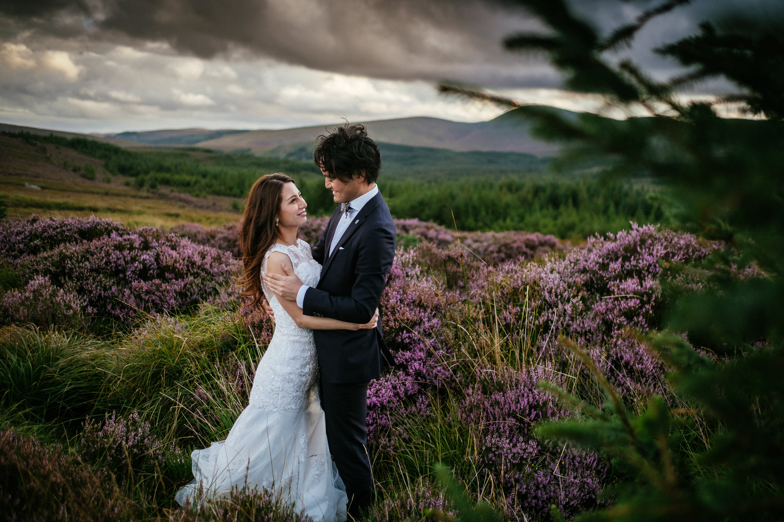 Eloping in Ireland 1 A bride and groom stand embracing in a field of purple heather, with rolling hills and a cloudy sky in the background—capturing a timeless moment of wedding photography when eloping in Ireland.