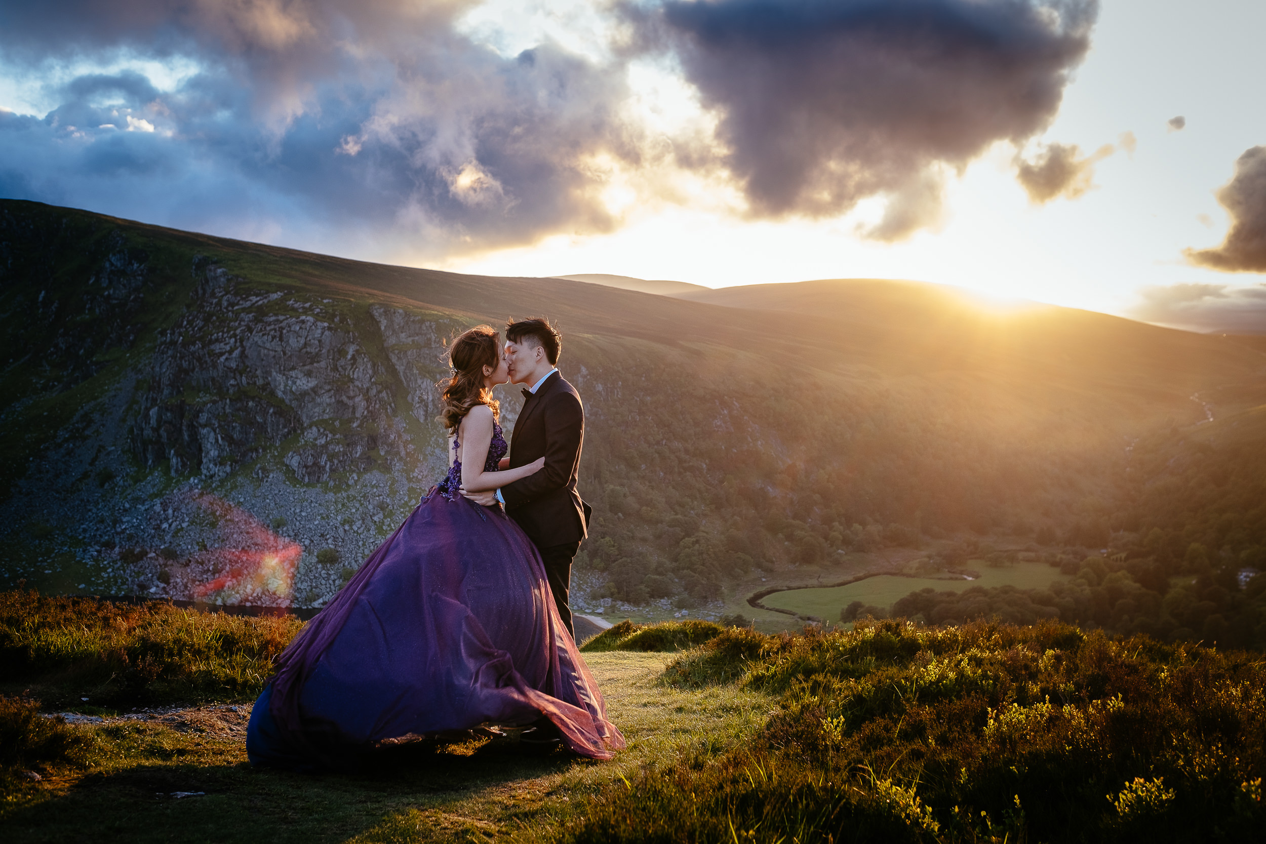 A couple in formal attire embraces on a grassy hilltop at sunset, experiencing the magic of eloping in Ireland, with rolling hills and dramatic clouds painting a breathtaking backdrop.