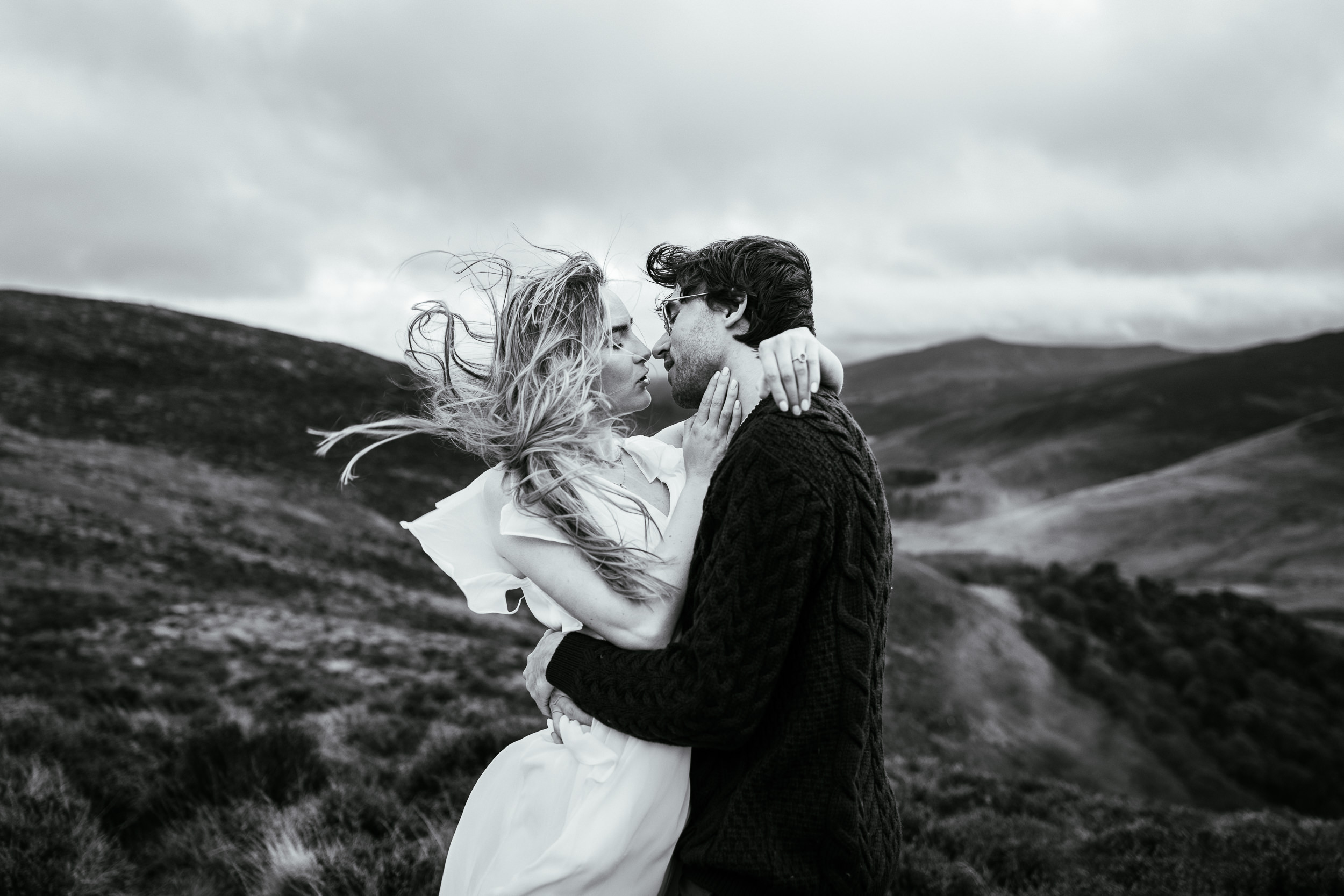 Engagement Shoot 16 A couple stands together on a grassy hillside with a lake and mountains in the background as the sun sets.