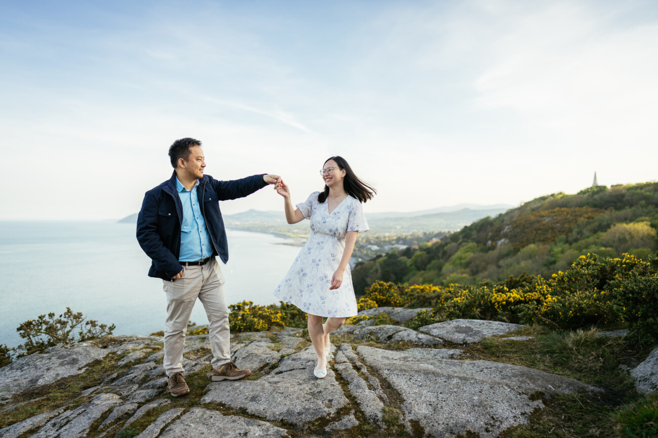 A man and woman hold hands and smile while standing on a rocky hilltop with the sea and distant landscape in the background.