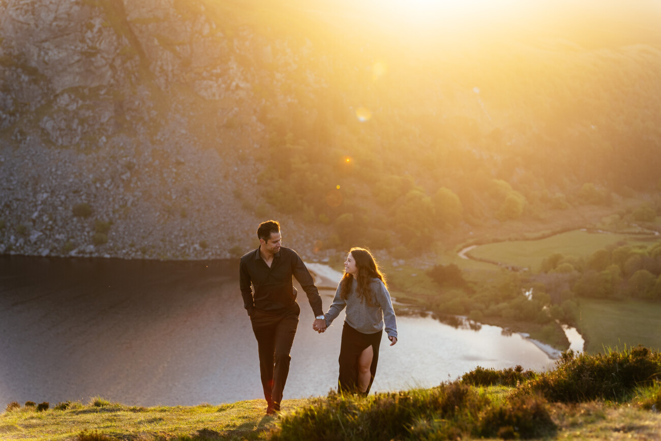 A man and woman hold hands and walk up a grassy hill near a lake at sunset, with sunlight illuminating the landscape.