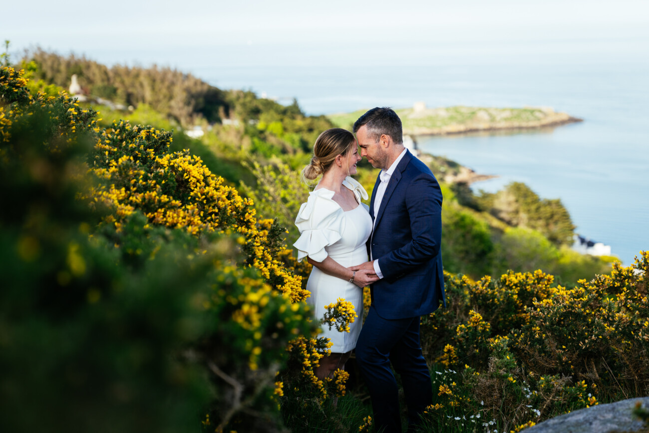 A couple stands close together in a field of yellow flowers on a hillside overlooking the sea, with greenery and coastal landscape in the background.