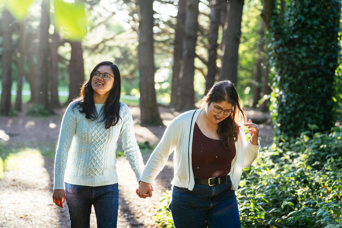 Two people walk hand in hand along a sunlit forest path, both wearing glasses and casual clothing, surrounded by trees and greenery.