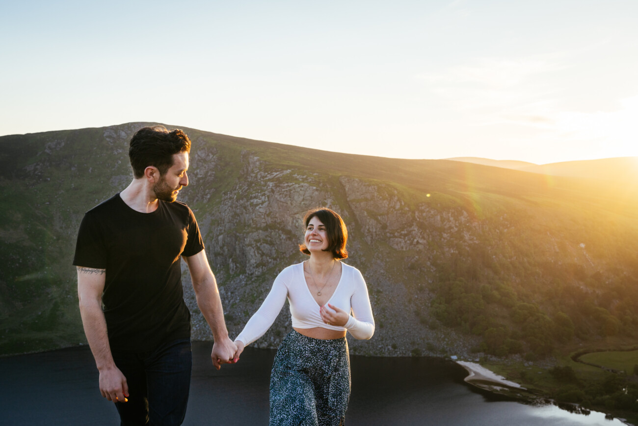 A couple holding hands and walking outdoors near a lake and hills at sunset, with sunlight illuminating the scene.