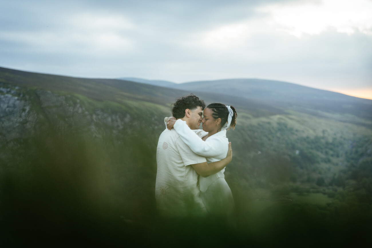 Two people embrace and kiss outdoors on a hillside with green mountains and a cloudy sky in the background.