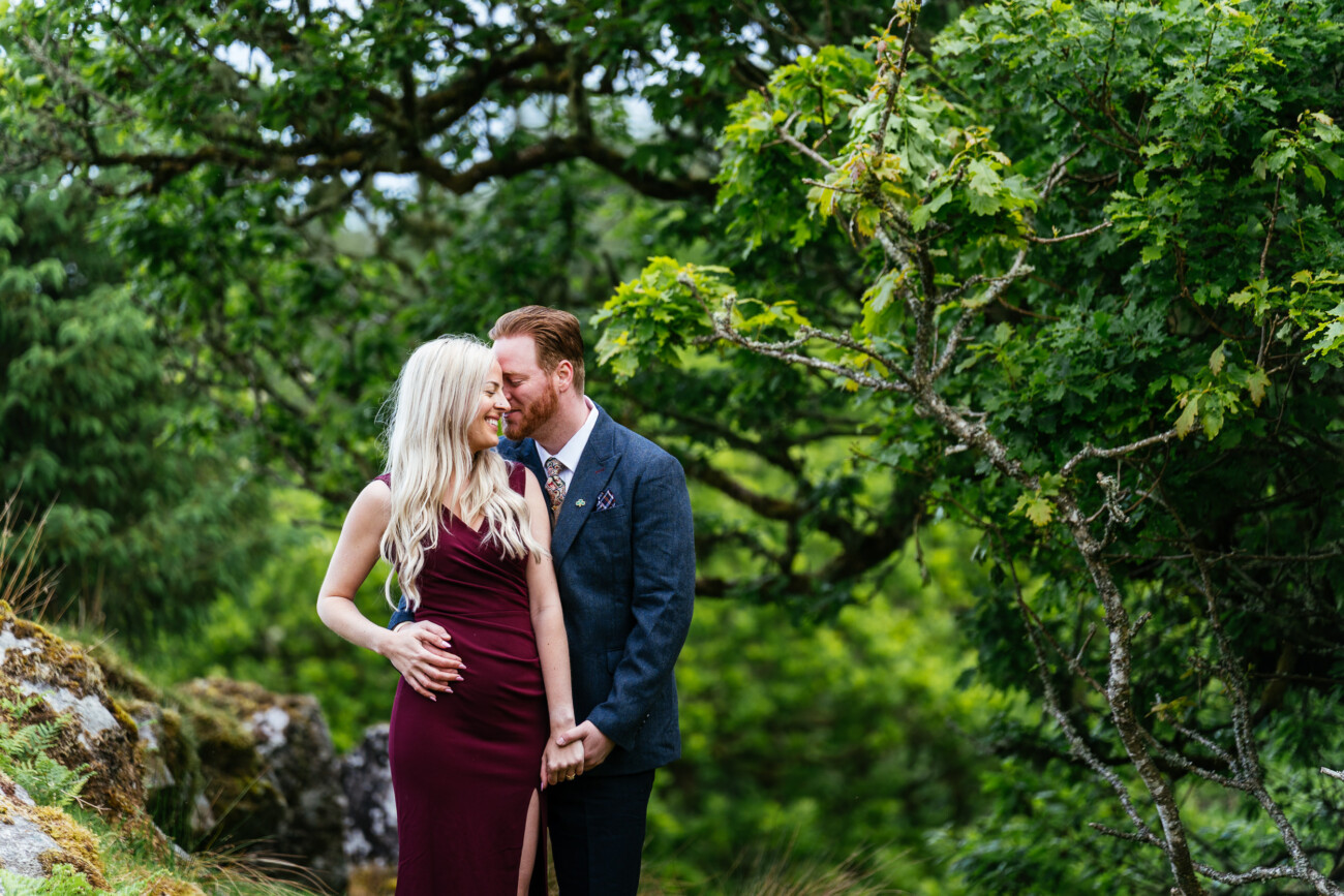 A couple stands close together in a lush, green outdoor setting; the man gently kisses the woman's temple as they hold hands and smile.