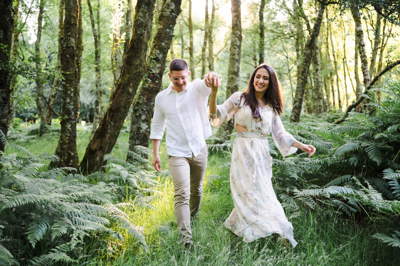 A man and woman, both smiling, walk hand in hand through a lush, green forest with tall trees and ferns.