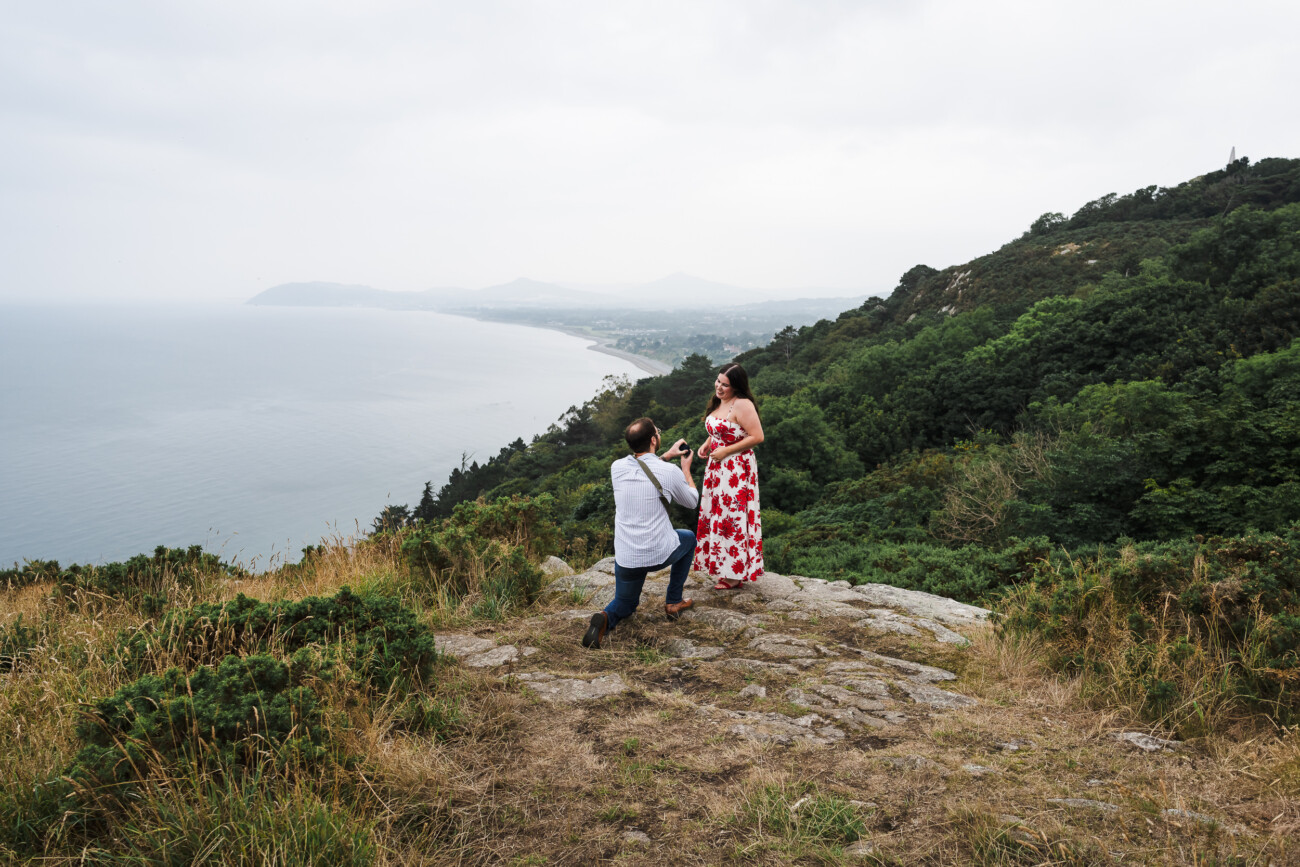 A man kneels and proposes to a woman in a red floral dress on a rocky hilltop overlooking the ocean and coastline, surrounded by greenery.