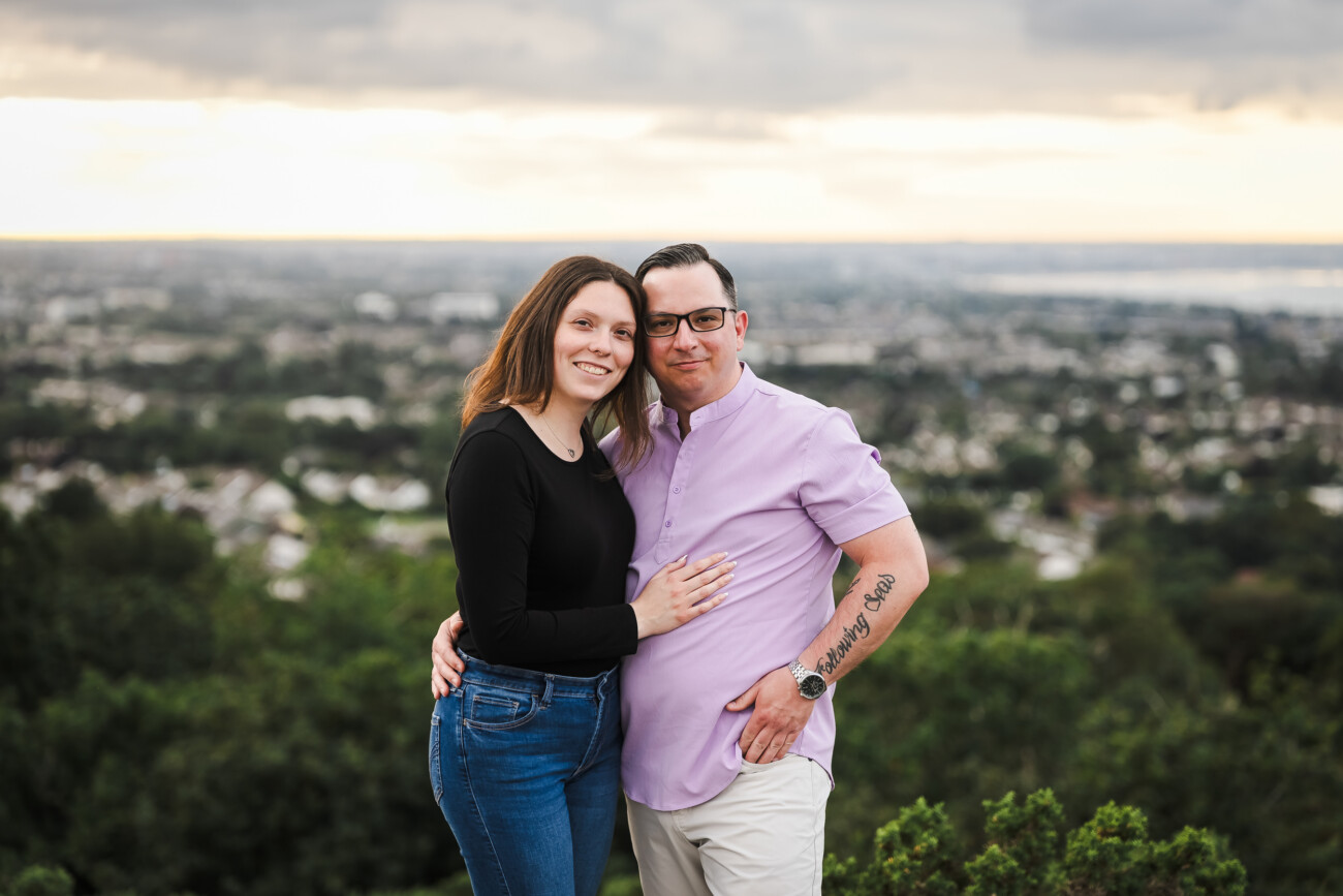A couple stands close together outdoors, smiling at the camera with a cityscape and cloudy sky in the background.