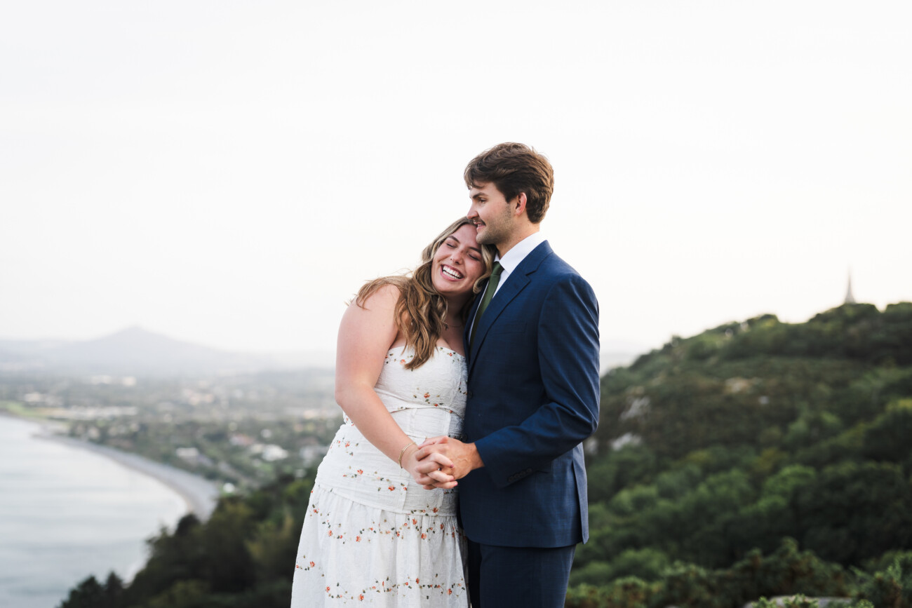 A couple dressed in formal attire stands close together, smiling and holding hands, with a scenic landscape of hills and water in the background.