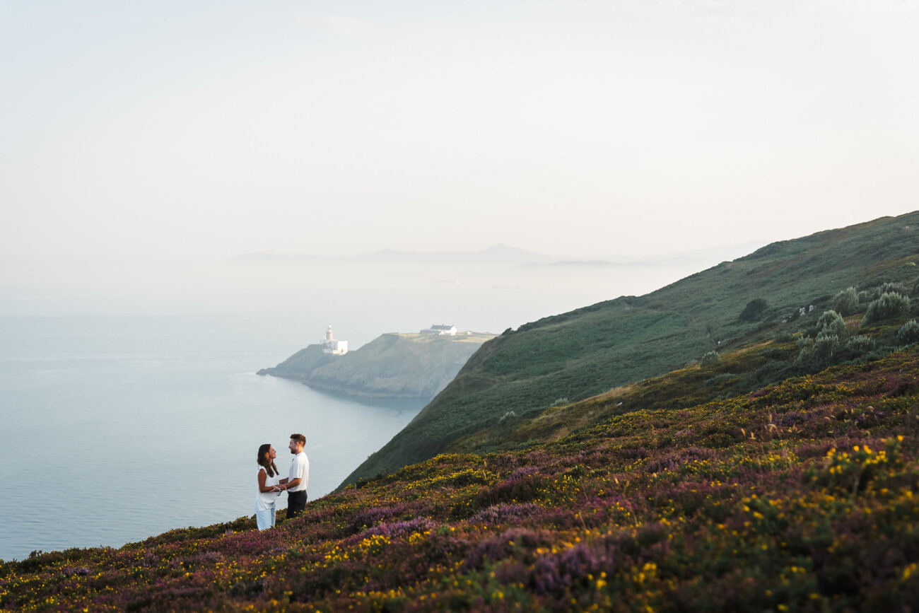 A couple stands on a hillside covered in wildflowers, overlooking the sea and a distant lighthouse on a coastal promontory.