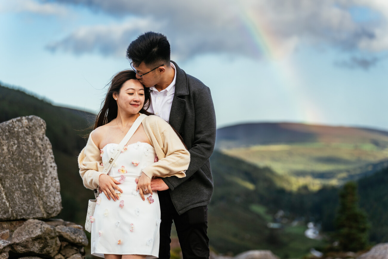 A couple stands outdoors with a rainbow and hills in the background; the man embraces and kisses the woman's head while she smiles with her eyes closed.