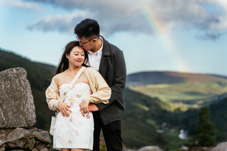 Ireland Travel Tips for US Visitors: What to Know Before You Go 1 A couple stands outdoors with a rainbow and hills in the background; the man embraces and kisses the woman's head while she smiles with her eyes closed.