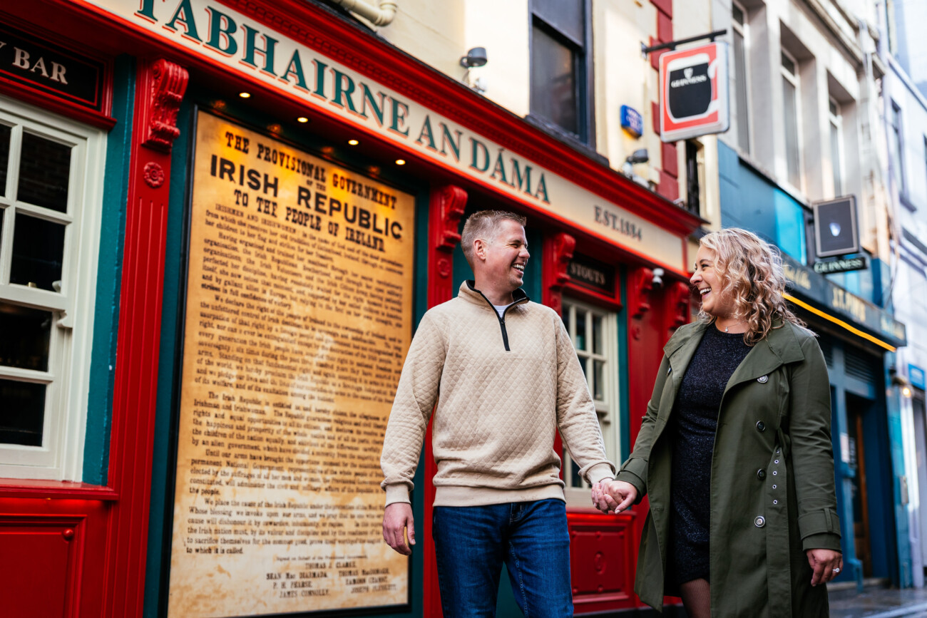 A man and woman holding hands and smiling stand outside a red-fronted Irish pub with a large Proclamation of the Irish Republic in the window.