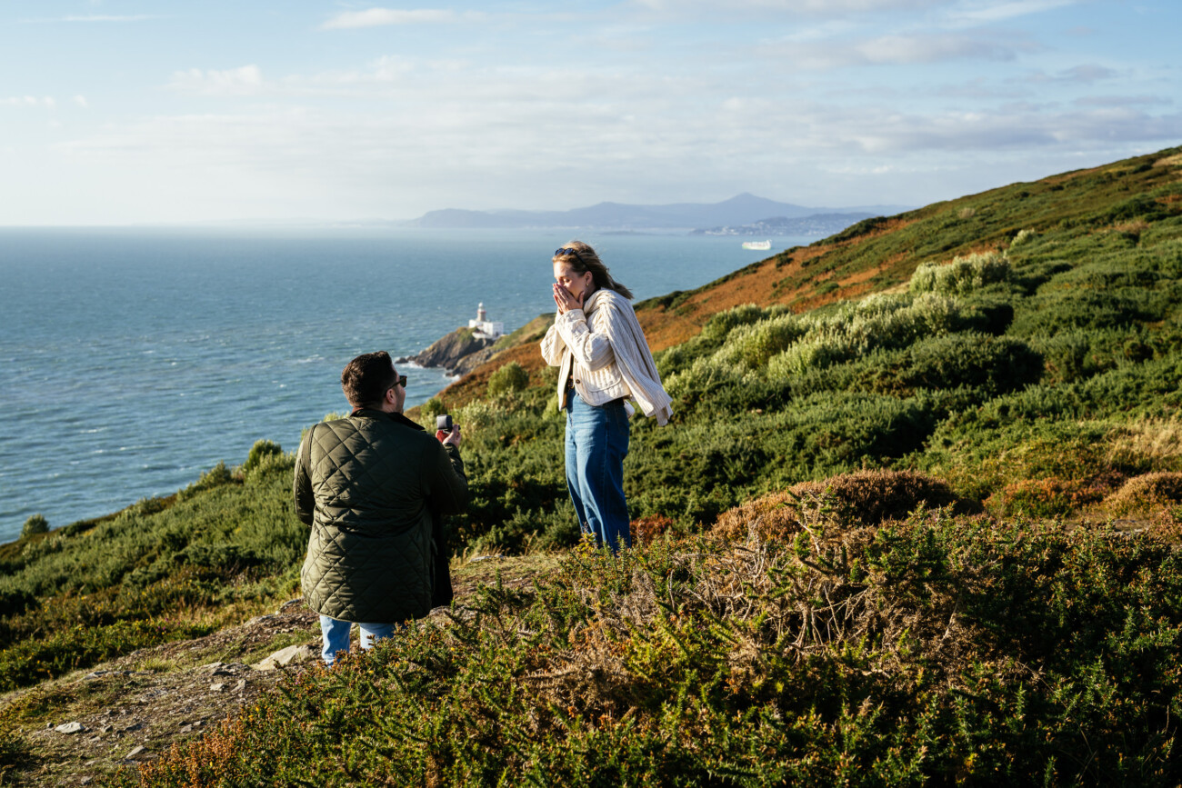A person kneels and presents a ring to another person on a grassy hillside overlooking the ocean, with a lighthouse and mountains in the background.