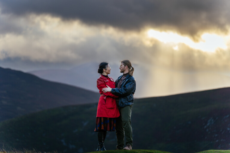 A couple stands facing each other on a grassy hill, with dramatic clouds and sun rays breaking through in the background.