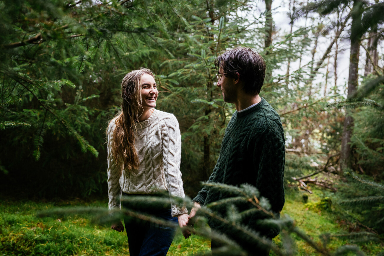 Two people wearing sweaters hold hands and smile at each other while walking through a forest with evergreen trees.