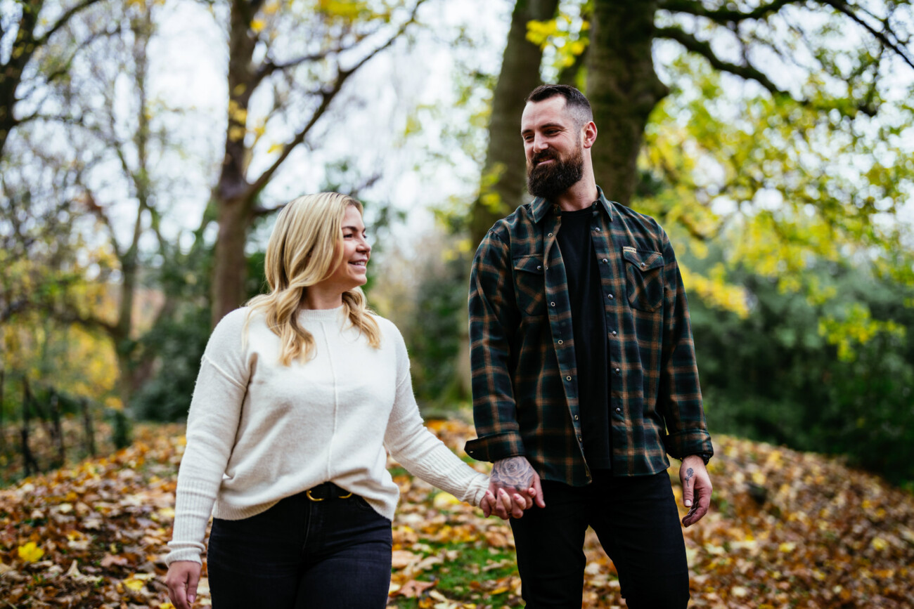 A man and a woman walk hand in hand through a park with fallen leaves and trees in the background, both smiling at each other.
