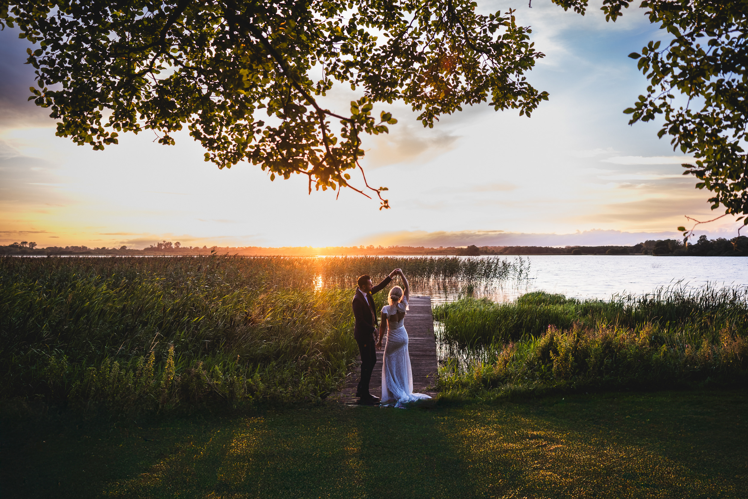 A bride and groom dance on a dock by a lake at sunset, surrounded by tall grass and trees, beautifully captured by a documentary wedding photographer in Ireland.