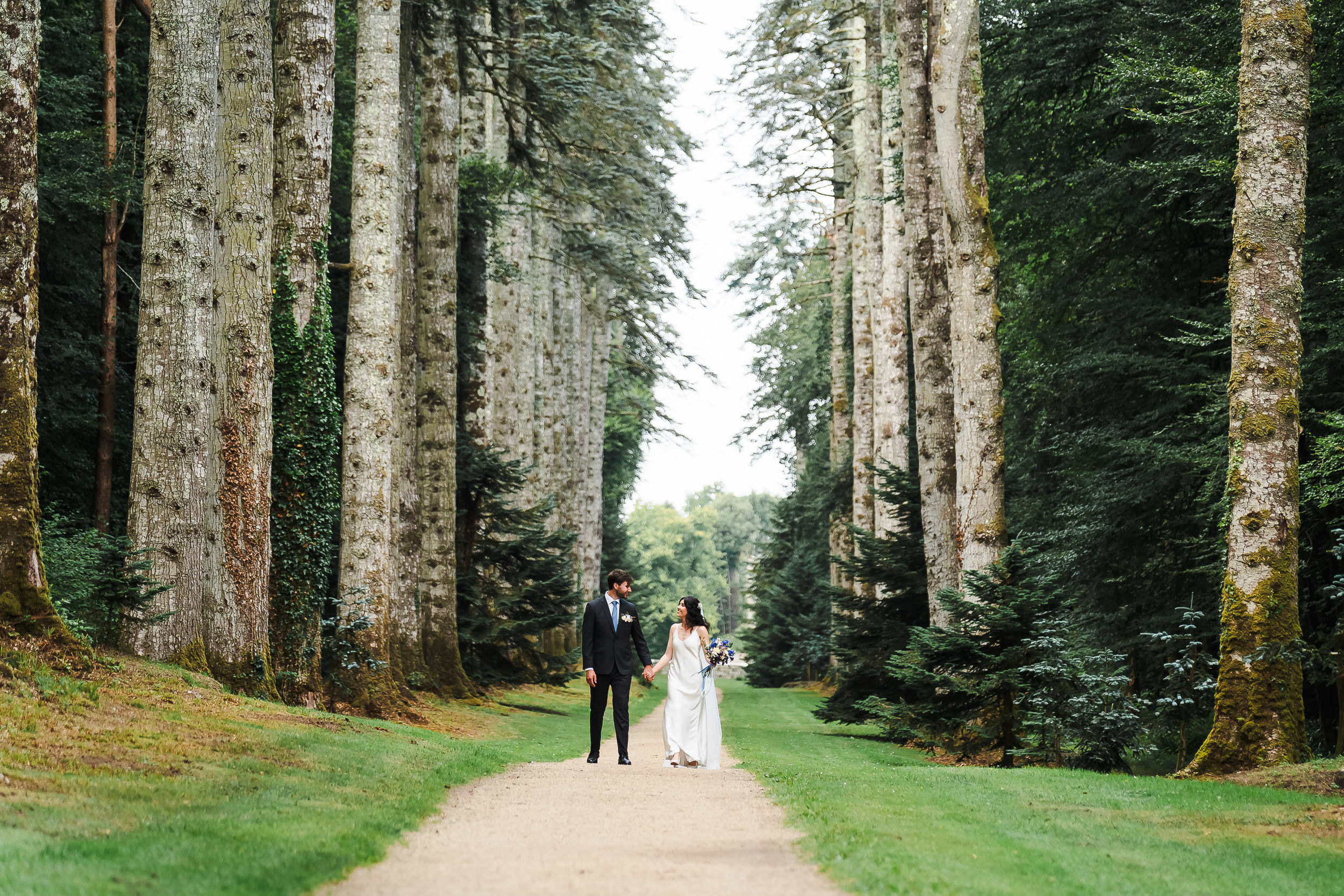 A bride and groom walk hand in hand down a tree-lined path in a lush, green forest, capturing a timeless moment of wedding photography.