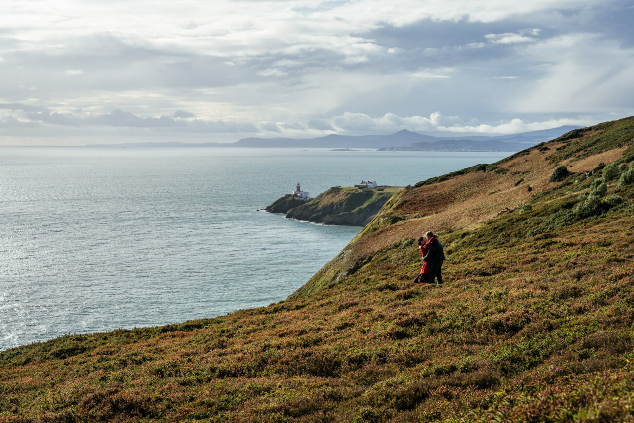 Two people stand on a grassy hillside overlooking the ocean with a lighthouse and distant mountains visible under a cloudy sky.