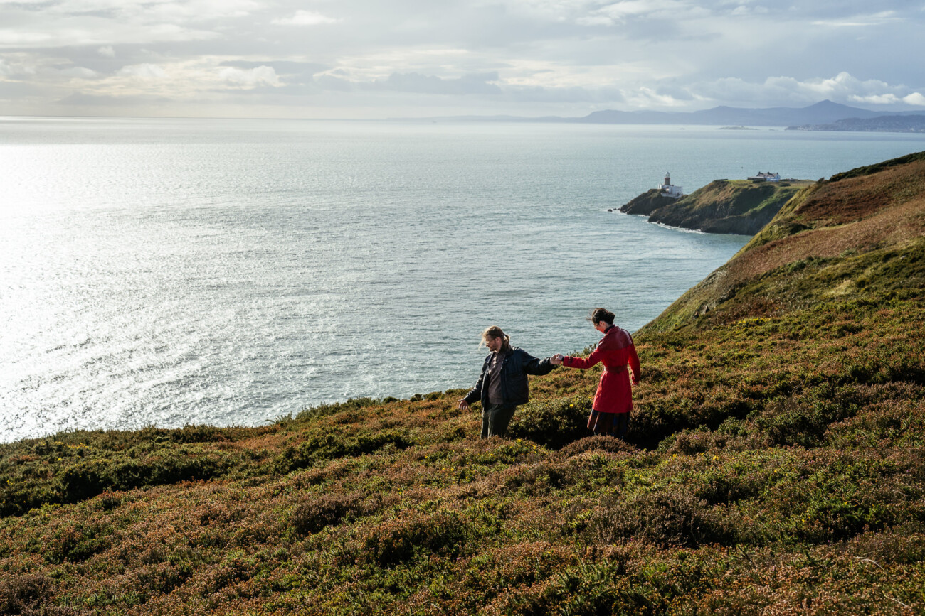 Two people walk hand in hand along a grassy hillside overlooking the sea, with a lighthouse visible in the distance under a cloudy sky.