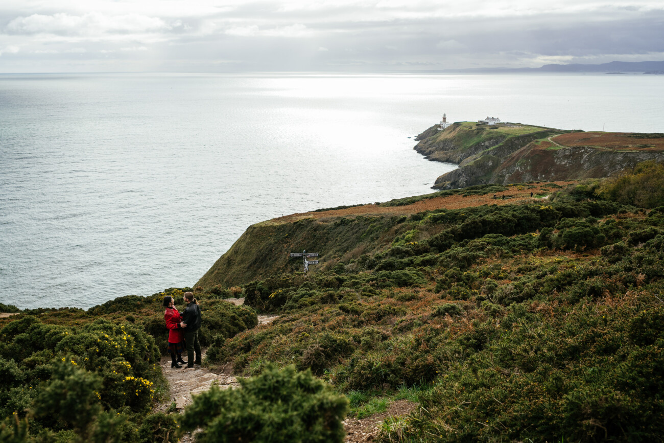 Two people stand on a coastal hillside path overlooking the sea, with cliffs, greenery, and a distant lighthouse under a cloudy sky.