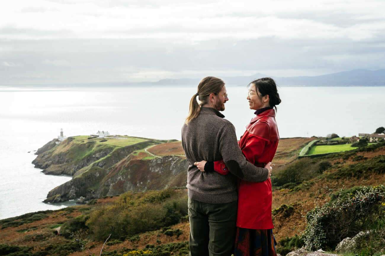 Couple standing on the edge of the Howth Head cliffs in Dublin, Ireland, overlooking the sea during an autumn honeymoon photo session.