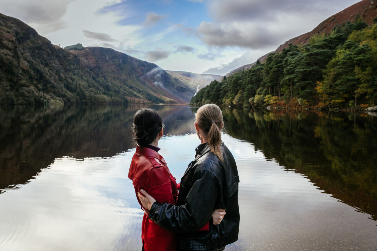 Two people wearing jackets stand close together at the edge of a calm lake, looking out at the surrounding hills and trees, with their backs to the camera.