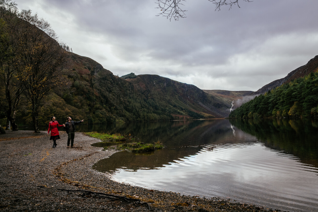 A couple walk along the shore of a calm lake in Glendalough Co Wicklow surrounded by forested hills and mountains, under a cloudy sky.