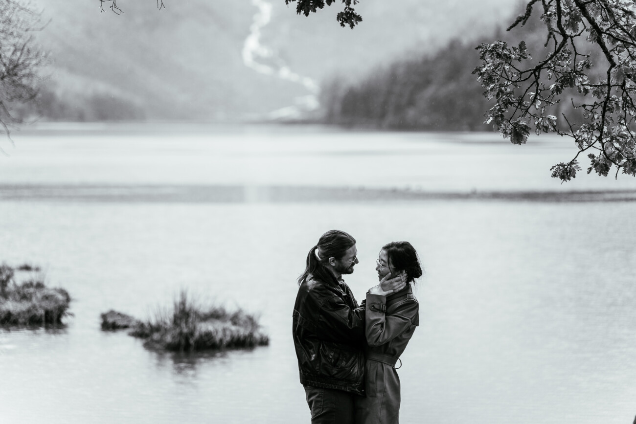 Two people stand close together by a lake, facing each other and touching faces, with trees and mountains visible in the background.