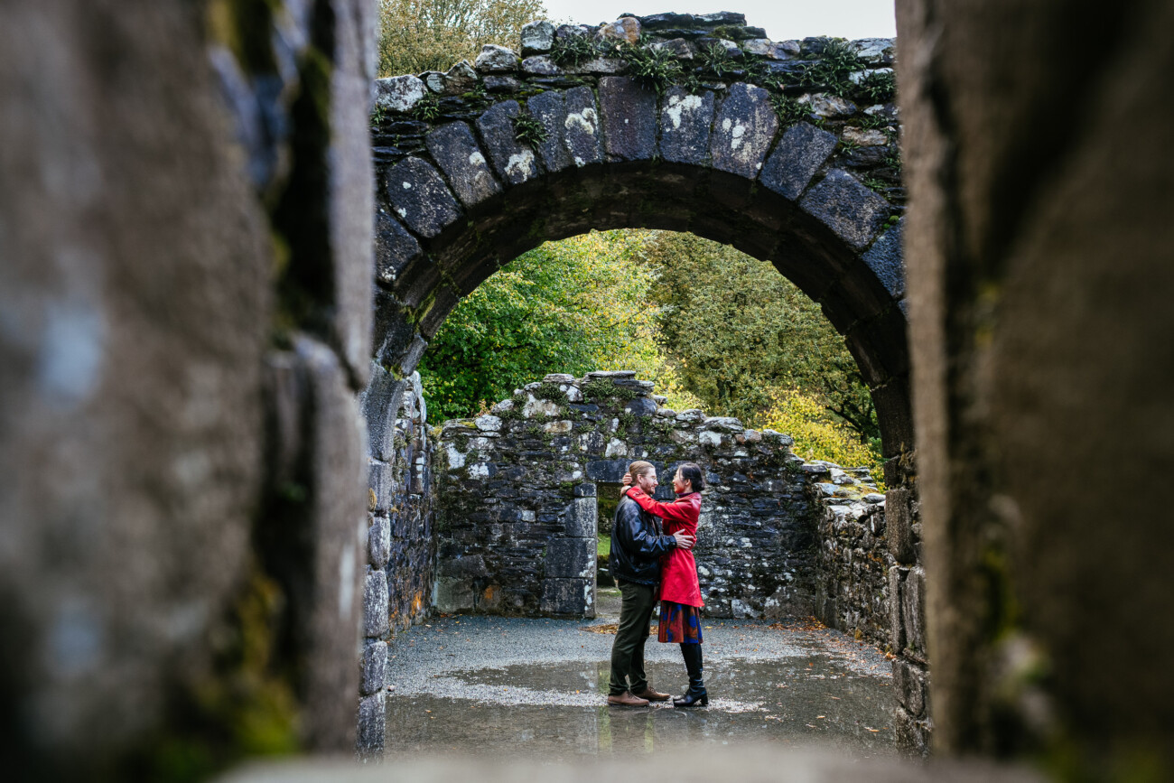 A couple stands close together under a stone archway amid the ruins of an old stone building, surrounded by greenery.
