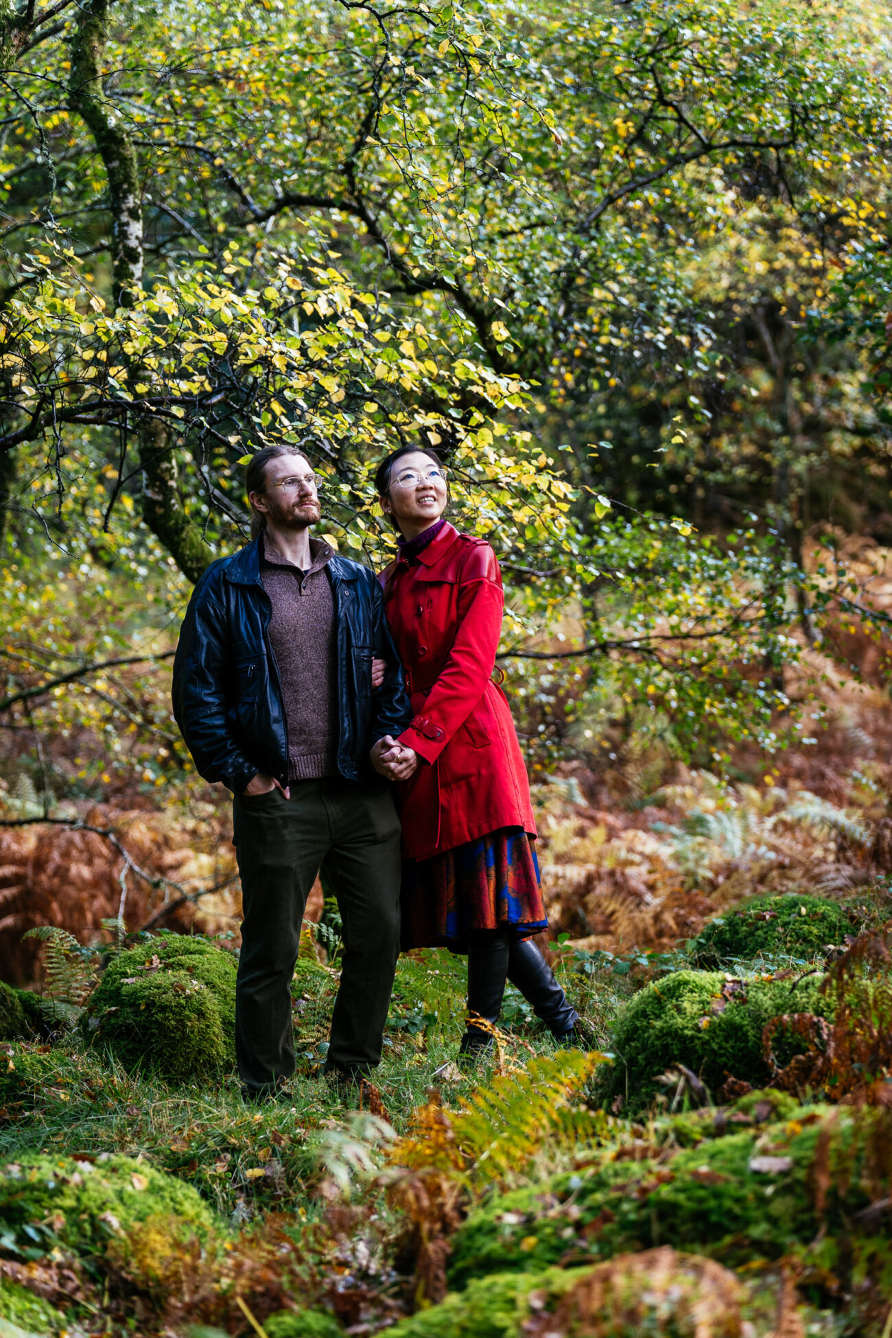 A man and a woman stand close together in a lush, green forest. The man wears a black jacket and the woman wears a bright red coat. Both look upward, surrounded by trees and foliage.