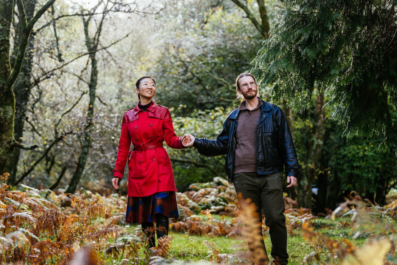 A woman in a red coat and a man in a black jacket hold hands while walking through a forested area with autumn foliage.