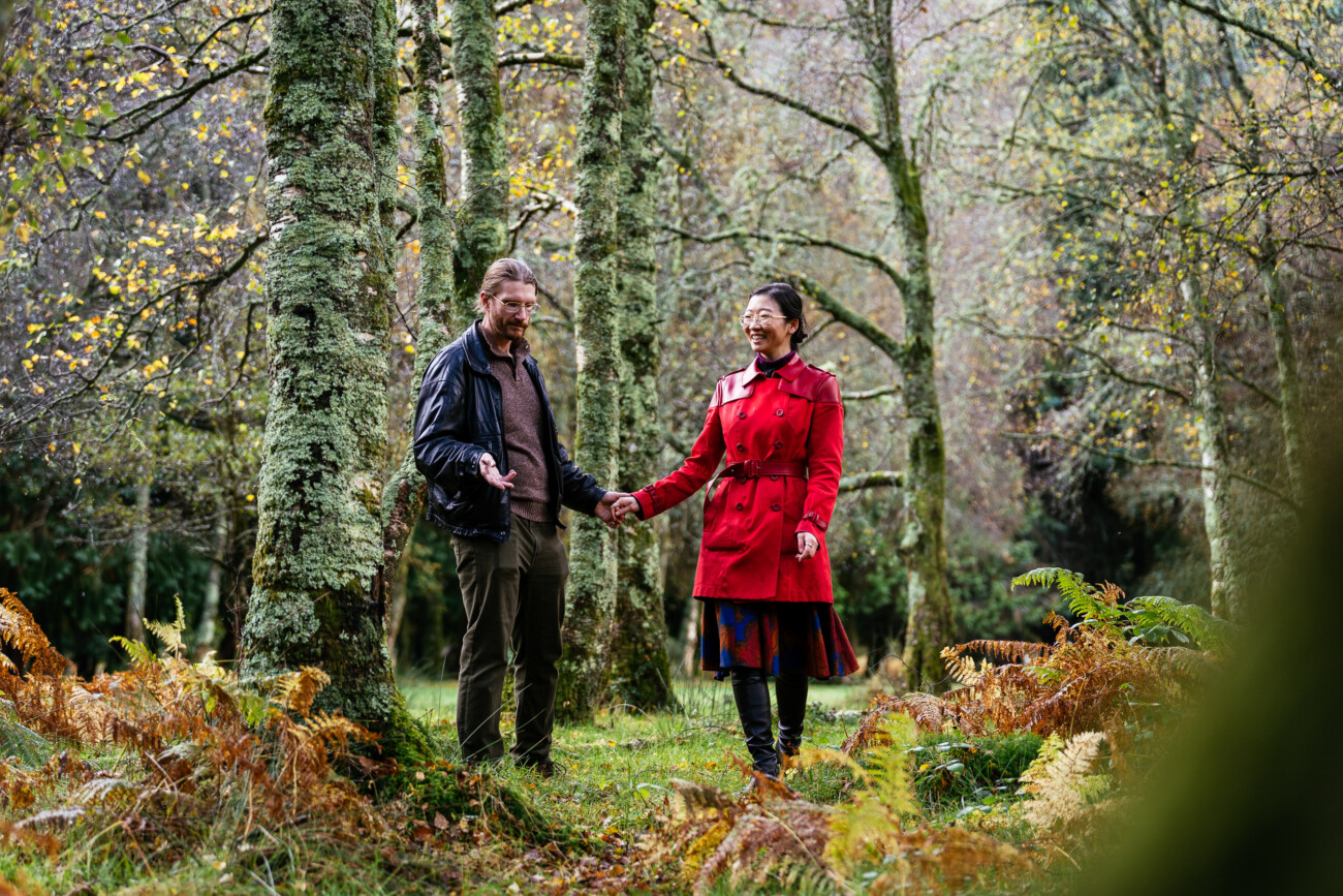 Two people stand holding hands in a forested area with green mossy trees and ferns on the ground, wearing jackets and autumn or winter clothing.