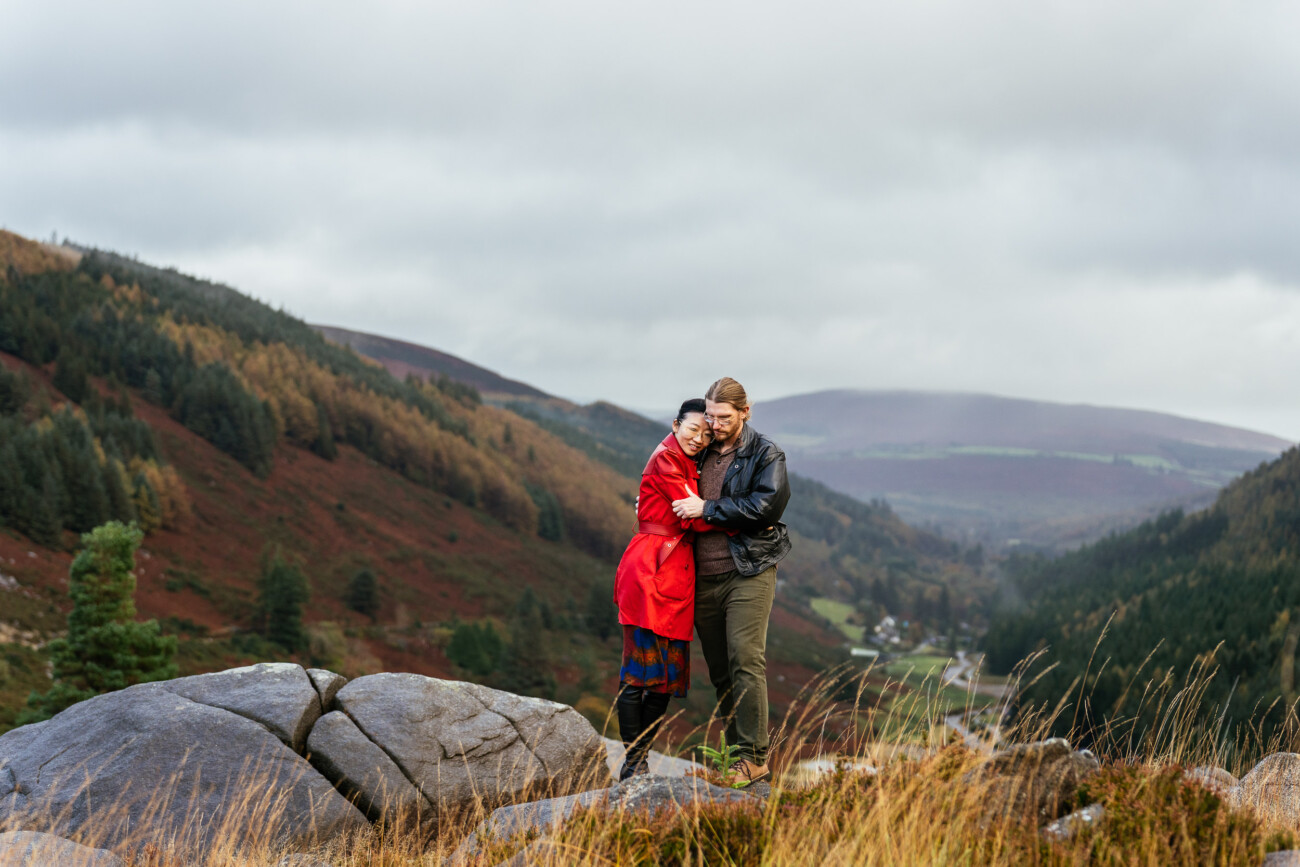 Two people stand on rocks in front of a valley with hills and trees, embracing each other. The sky is overcast and the landscape is covered in autumn colors.