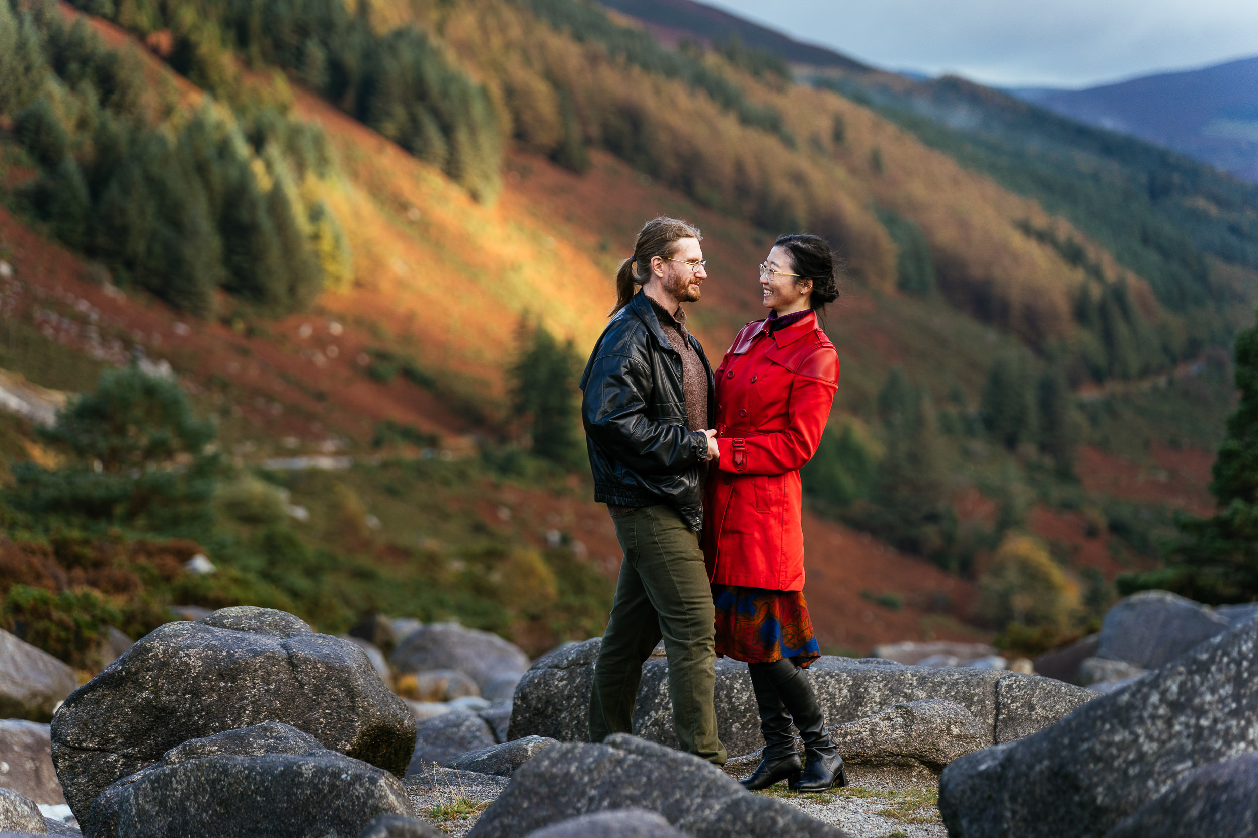 A couple stand on large rocks outdoors, facing each other and smiling, with a forested valley and hillside in the background during their couple shoot in Ireland. The lady is wearing a vibrant red coat which pops agains the dark background in a visually stunning image.