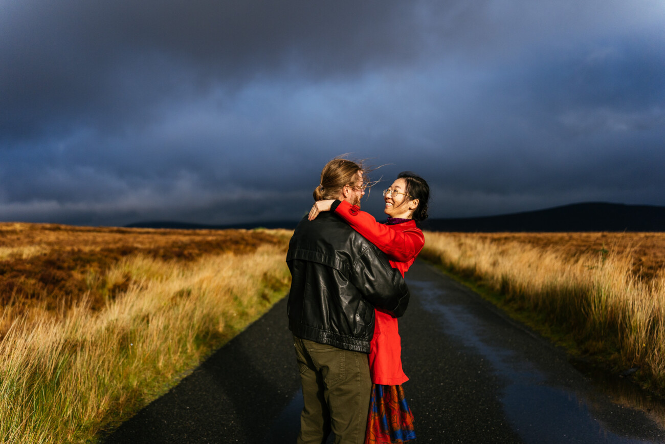 A couple stands embracing on a rural road surrounded by golden grass, under a dramatic cloudy sky.