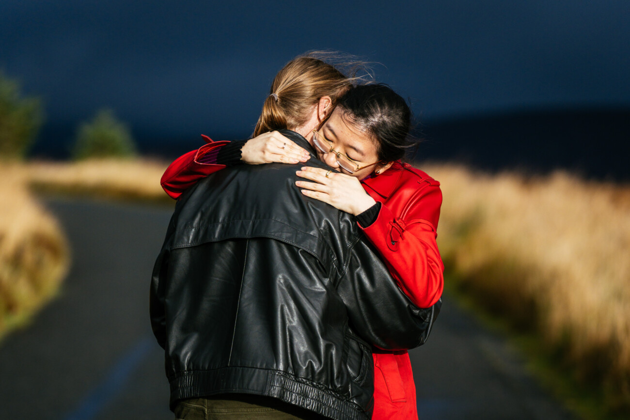 Two people embrace on an outdoor path, one wearing a black jacket and the other a red coat, with tall grass and a dark sky in the background.