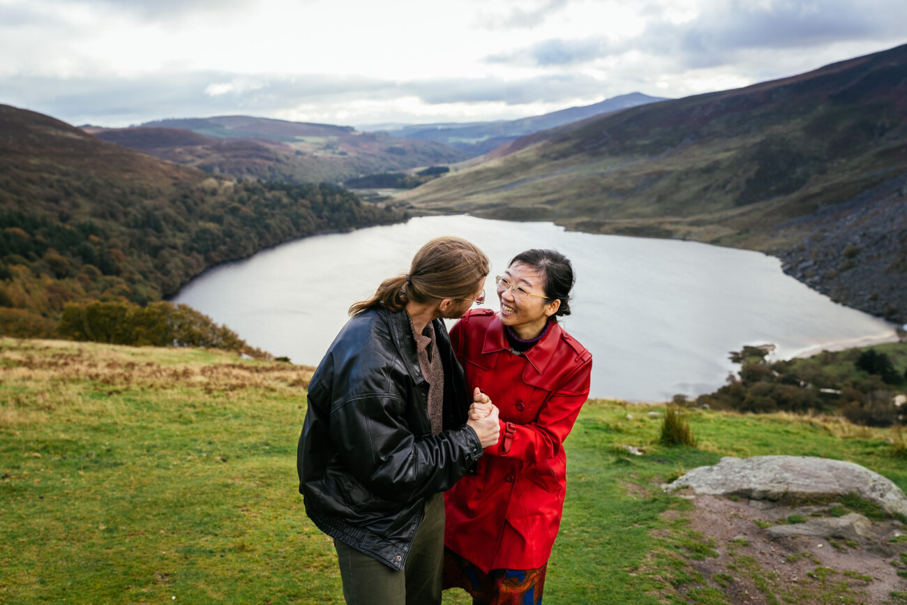Two people stand on grass above a lake and mountains, holding hands and smiling at each other. One wears a leather jacket, the other a red coat.