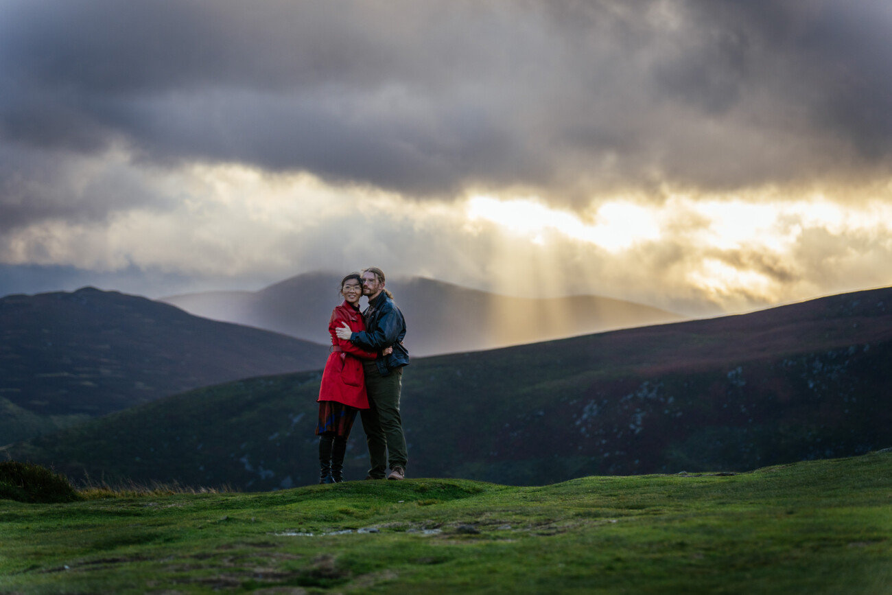 a coupleembrace on a grassy hill with mountains in the background and dramatic clouds with rays of sunlight breaking through at Guinness lake.