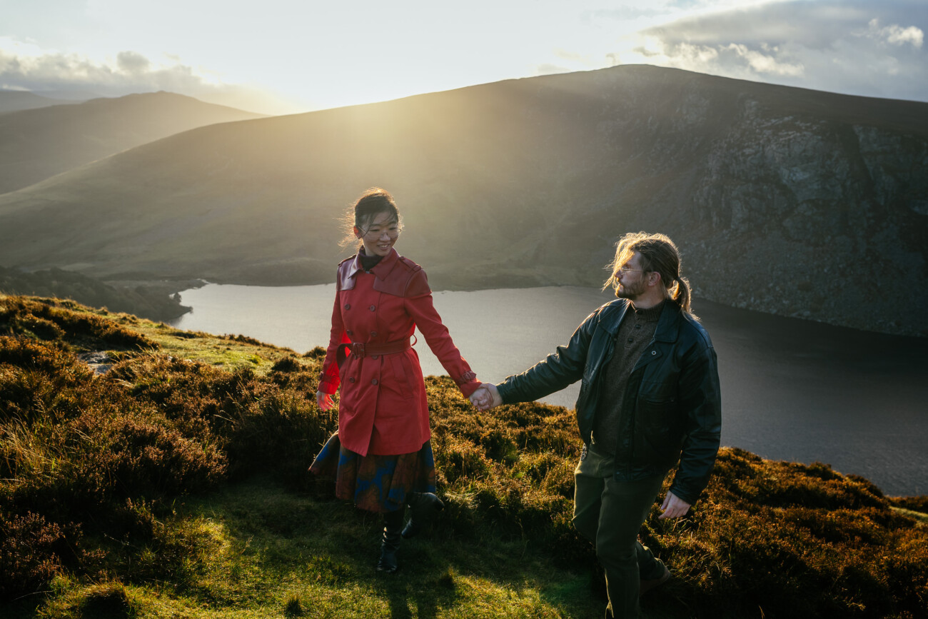 A woman in a red coat and a man walk hand in hand on a grassy hillside near a lake at sunset, with mountains in the background.