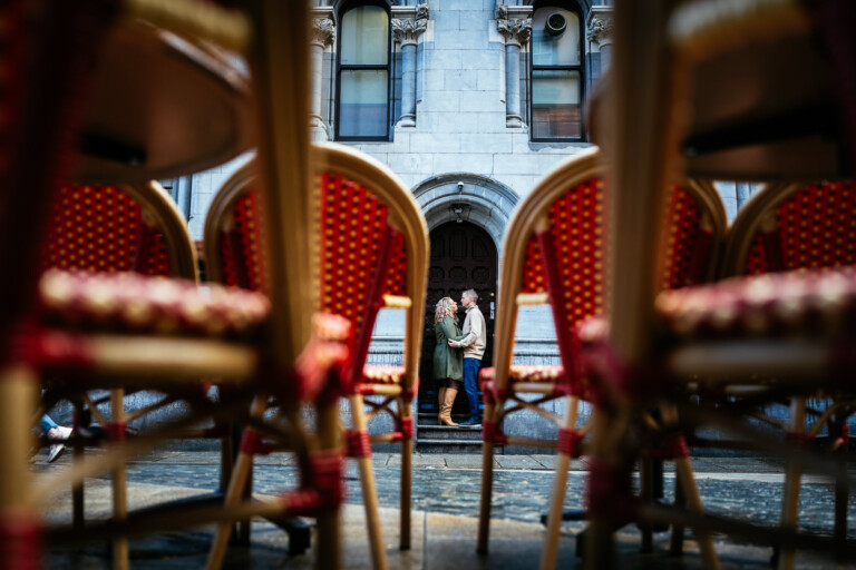 Two people embrace and kiss on a city street, framed through the legs of stacked red and yellow chairs, as a Dublin Engagement Photographer captures them against a backdrop of stone buildings with arched windows during their engagement shoot dublin temple bar.