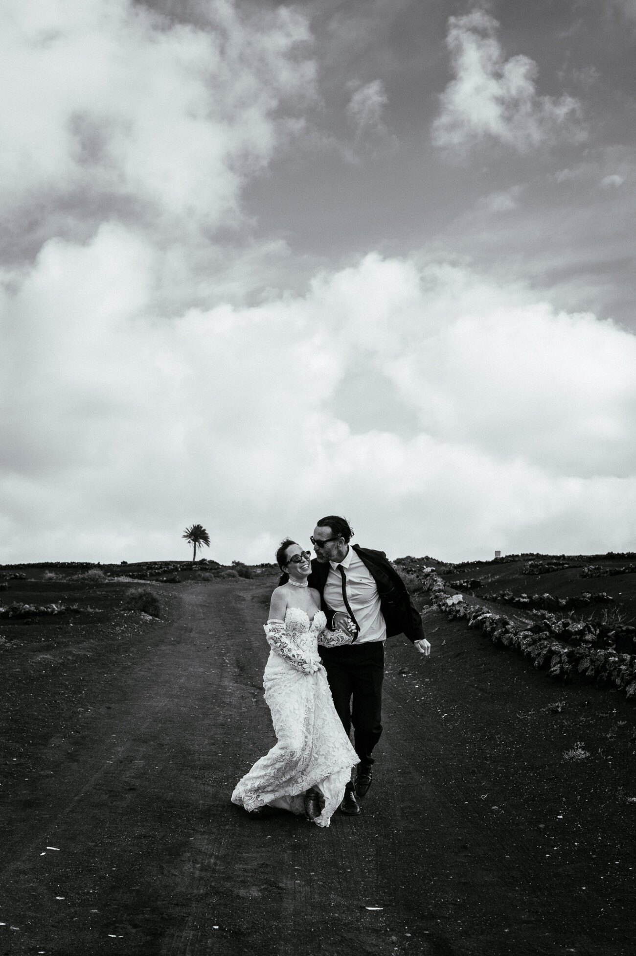 A bride and groom walk on a dirt path outdoors, holding each other and smiling, with a cloudy sky and open landscape in the background—perfect for destination wedding photography.