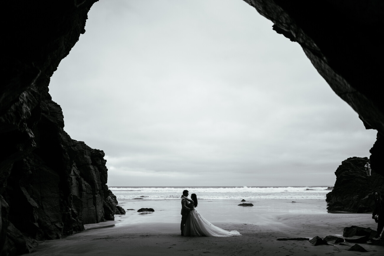 A couple in wedding attire stands embracing on a sandy beach, framed by the dark silhouette of a large cave opening—an unforgettable moment captured in destination wedding photography, with waves and a cloudy sky as their backdrop.