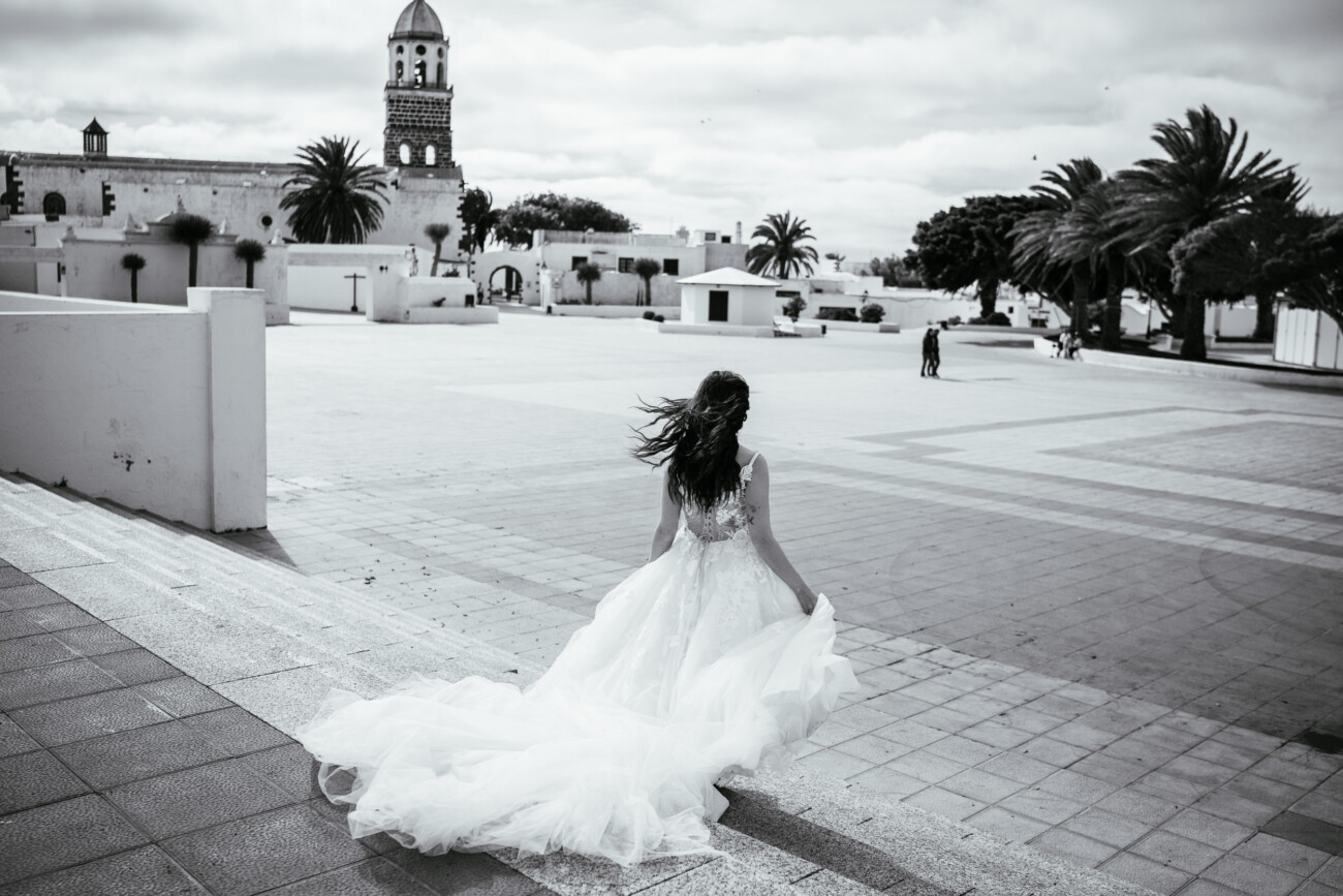 A woman in a wedding dress walks alone across a large, empty plaza with palm trees and a bell tower in the background, capturing the essence of destination wedding photography.