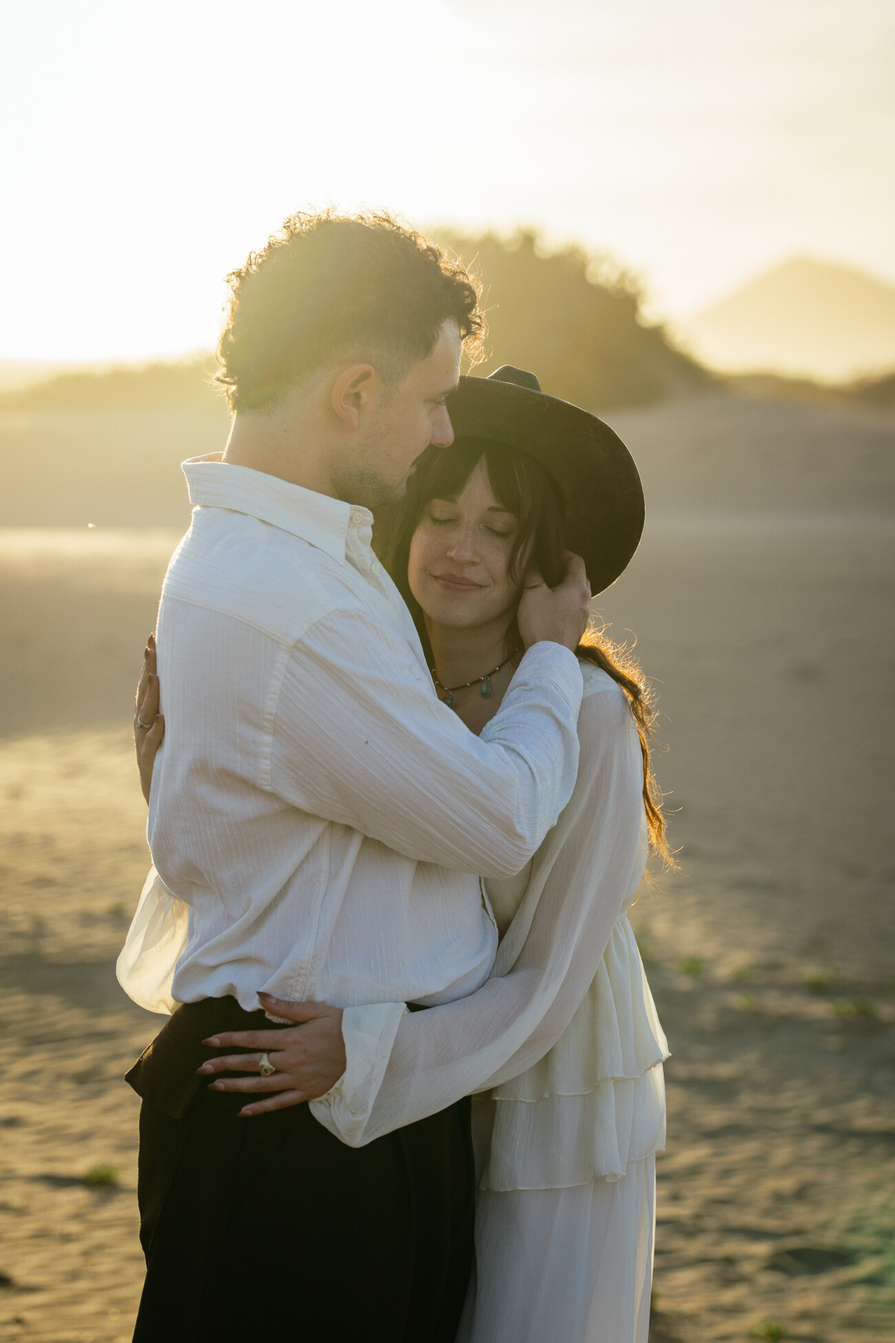 A couple in white clothing embraces on a sandy beach at sunset; the woman, wearing a black hat and smiling with her eyes closed, captures the magic of destination wedding photography.