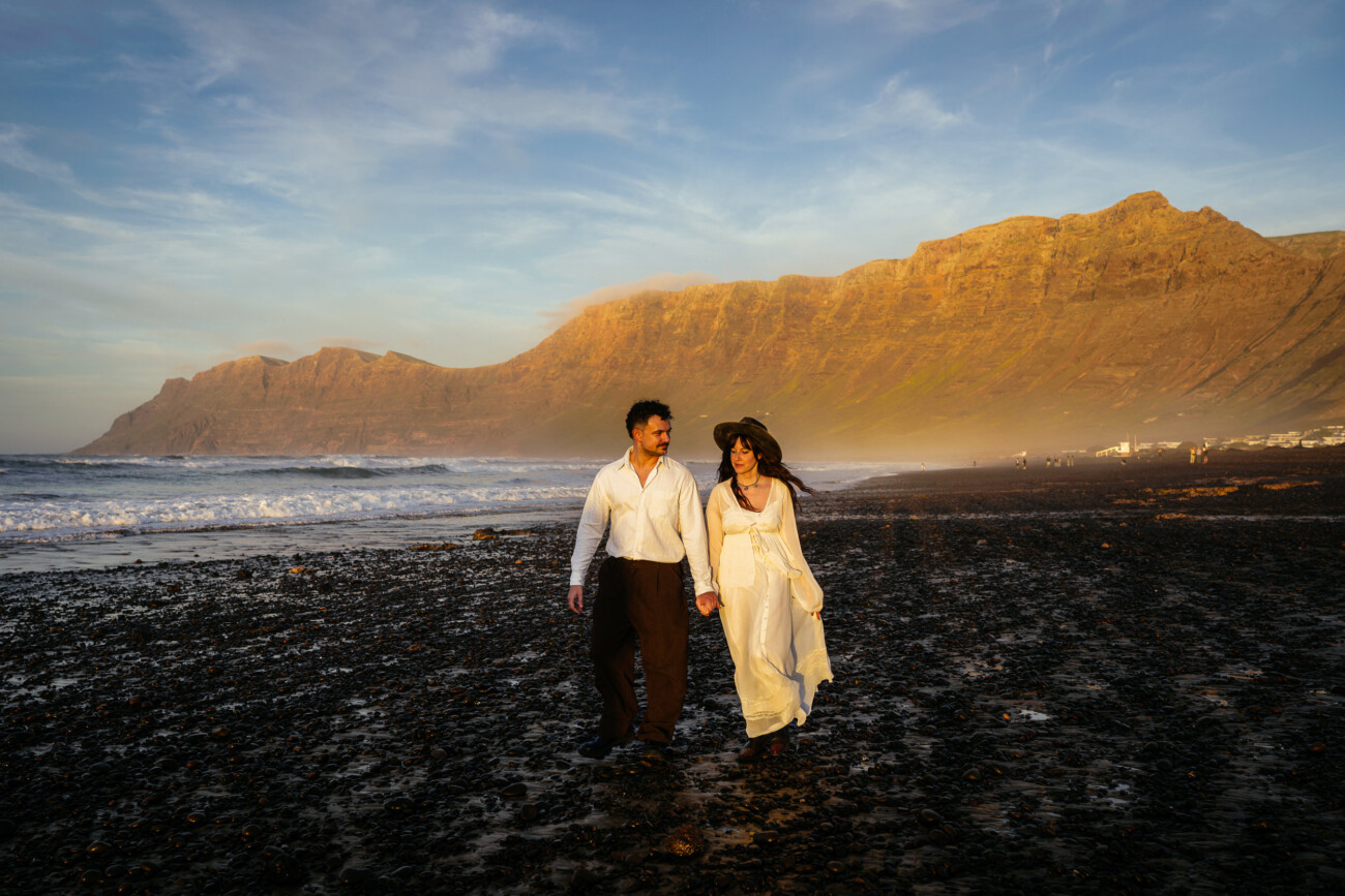 A man and woman walk hand-in-hand along a rocky beach at sunset, with mountains in the background—an idyllic moment perfect for destination wedding photography.