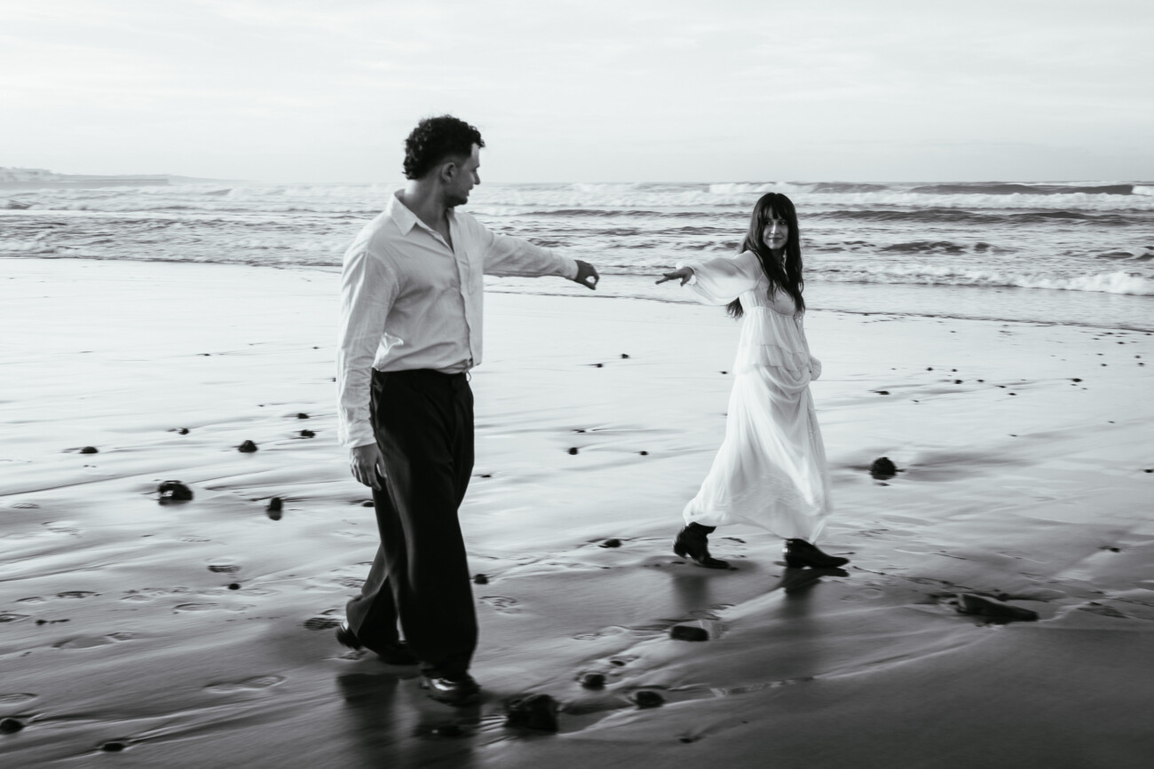 A man and woman walk on a sandy beach, reaching towards each other. Captured in black and white, this destination wedding photography scene features waves and rocks in the background, with both wearing light-coloured clothing.