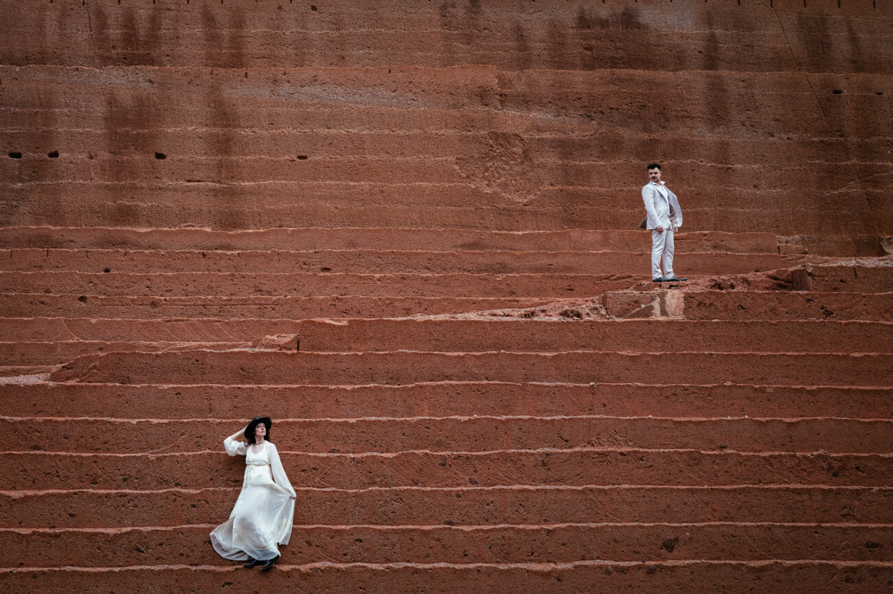 A woman in a white dress and a man in a white suit stand far apart on wide, stepped, reddish-brown stone terraces—a striking scene of destination wedding photography.